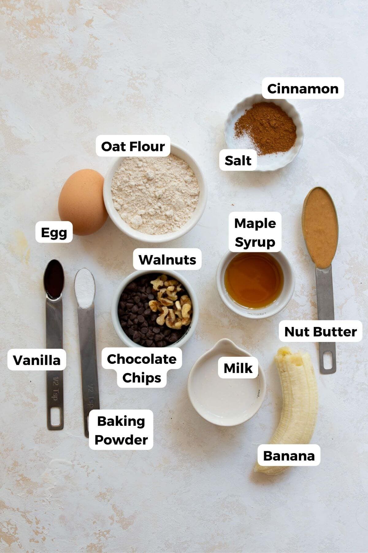Ingredients for healthy single serve banana bread laid out, including oat flour, banana, egg, nut butter, maple syrup, milk, walnuts, chocolate chips, baking powder, cinnamon, and salt.