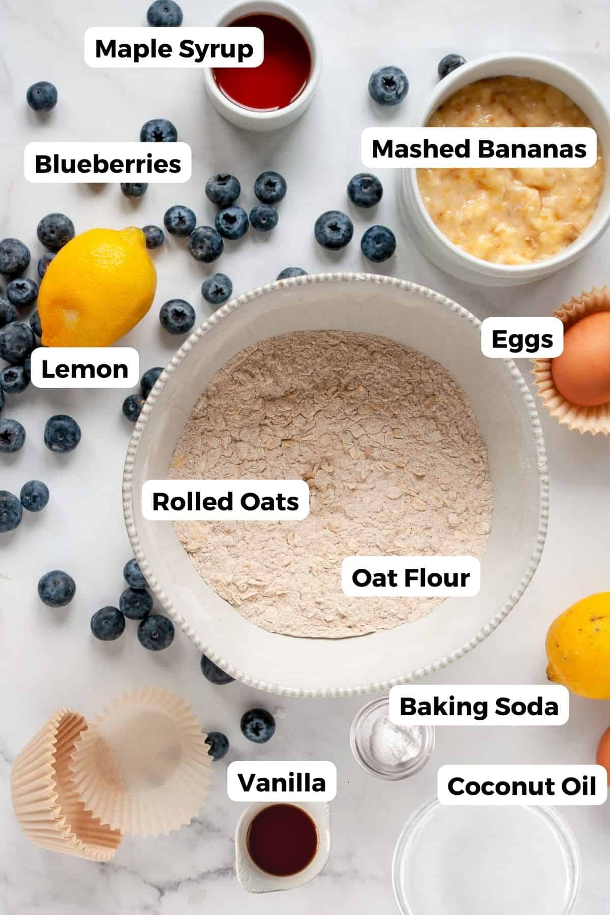 Ingredients for healthy banana blueberry oatmeal muffins including oat flour, rolled oats, blueberries, bananas, maple syrup, coconut oil, eggs and lemon.