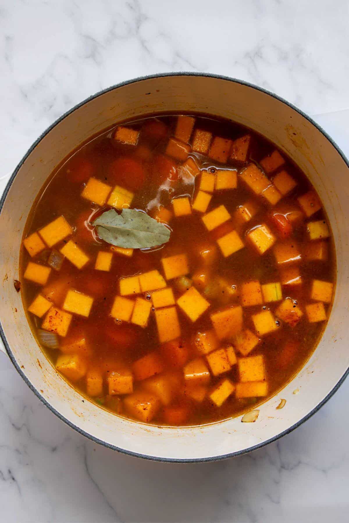 Bay leaf and vegetable broth being poured into a pot with butternut squash, carrots, and aromatics.