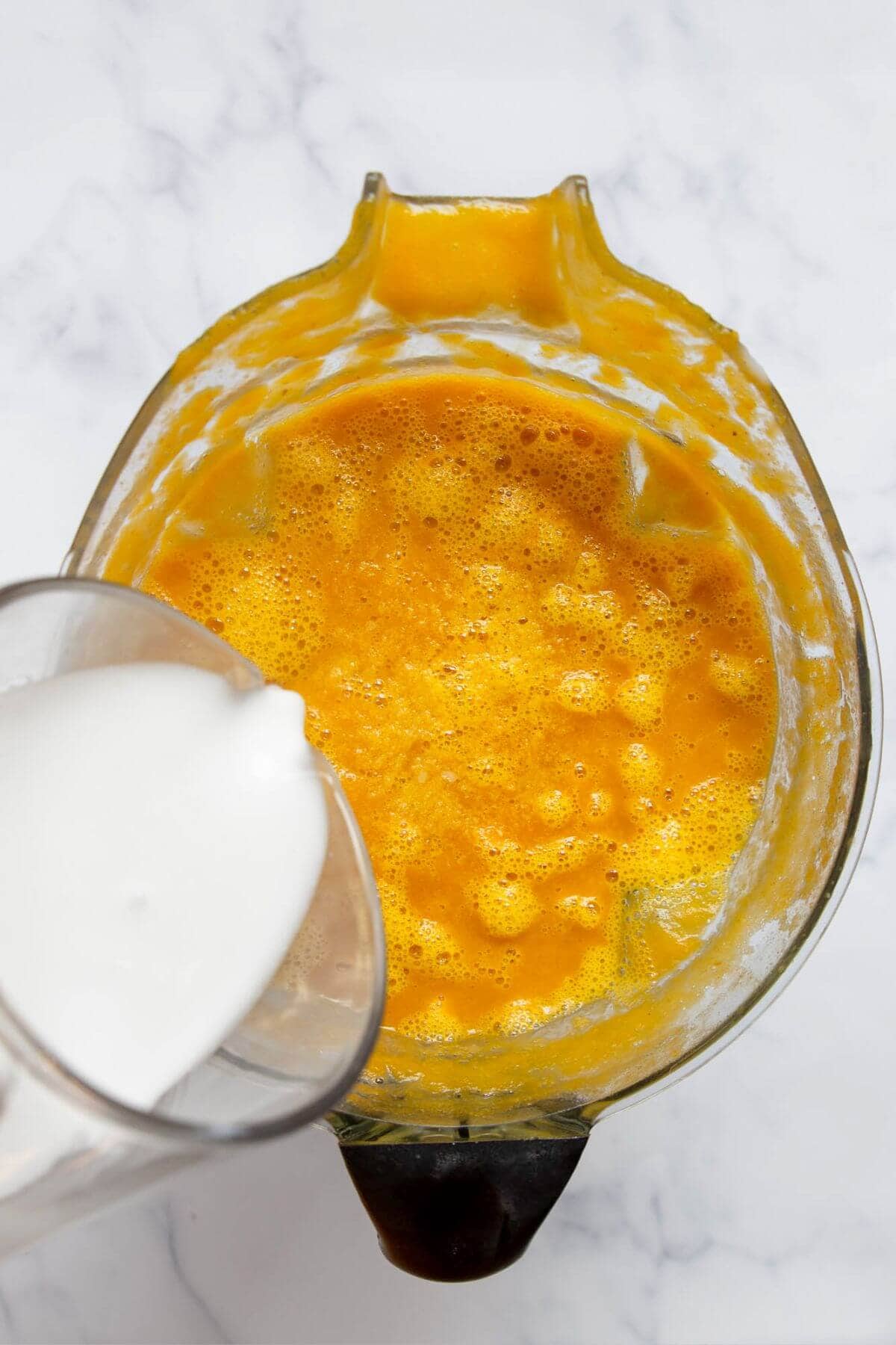 Pouring coconut milk into blended butternut squash and carrot soup to make it creamy and dairy-free.