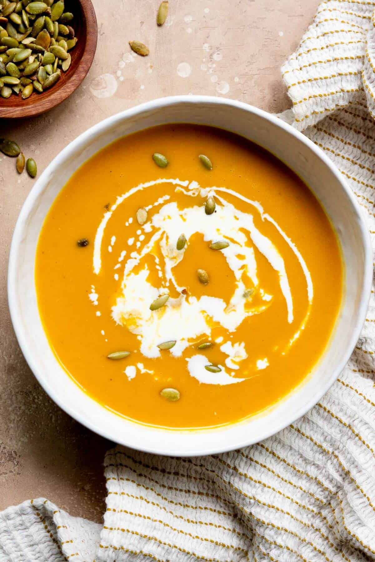 Creamy carrot and butternut squash soup served in a bowl, topped with a swirl of coconut milk and crunchy pumpkin seeds.
