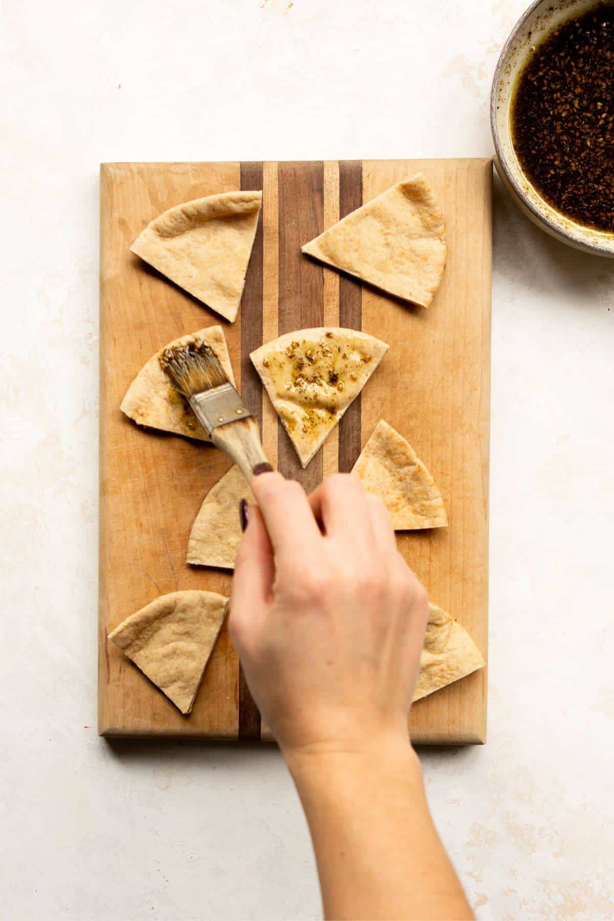 A hand brushing olive oil and za’atar seasoning onto cut pita bread triangles on a cutting board