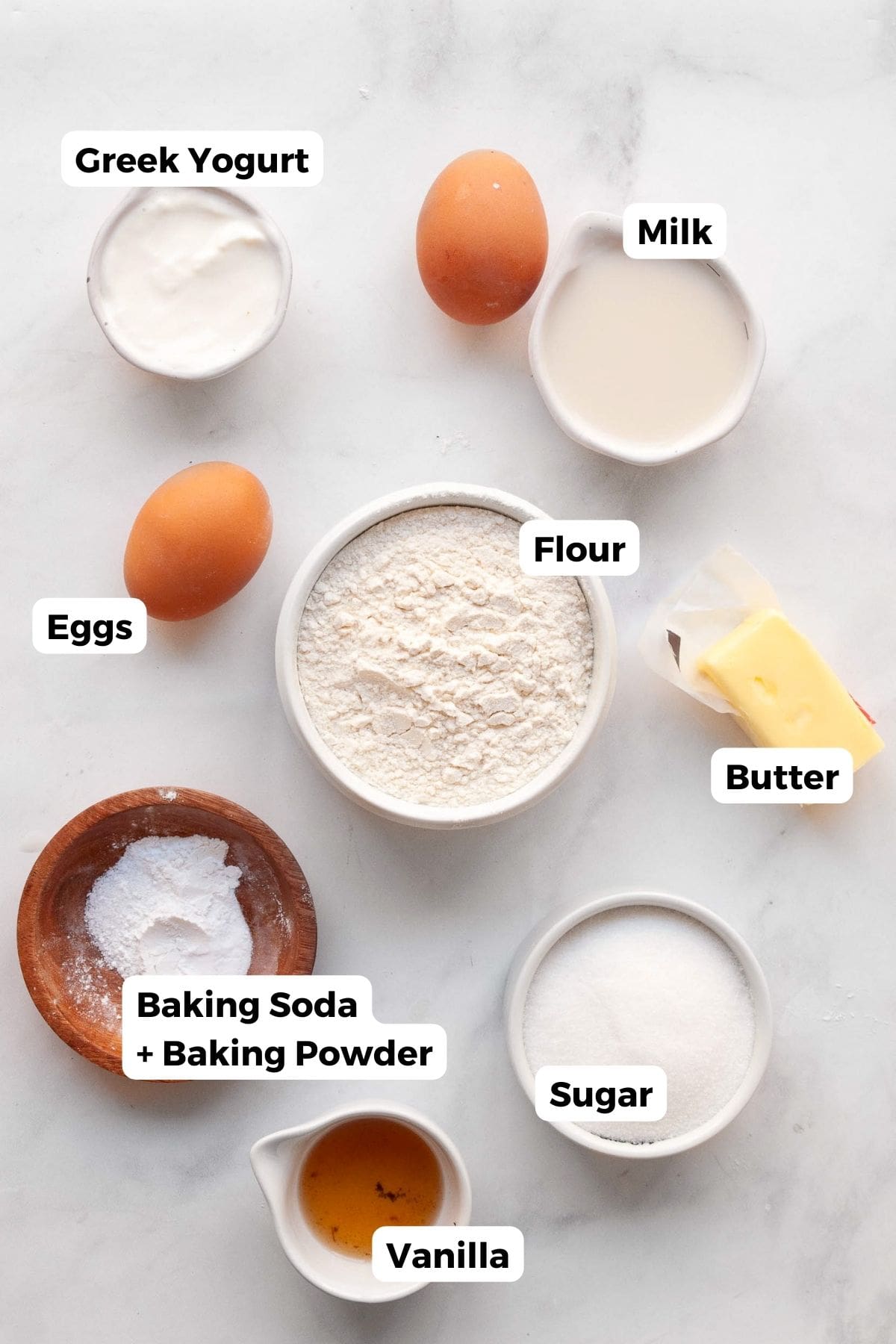 All ingredients in mini bundt cake recipe in small bowls with labels, including: flour, sugar, Greek yogurt, butter, vanilla, baking soda, baking powder, eggs, and milk.