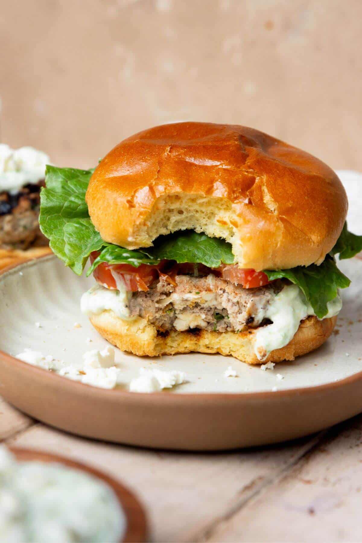 Bite taken out of a Greek turkey burger showing the juicy patty with melted feta inside, topped with tzatziki, tomato, and lettuce.