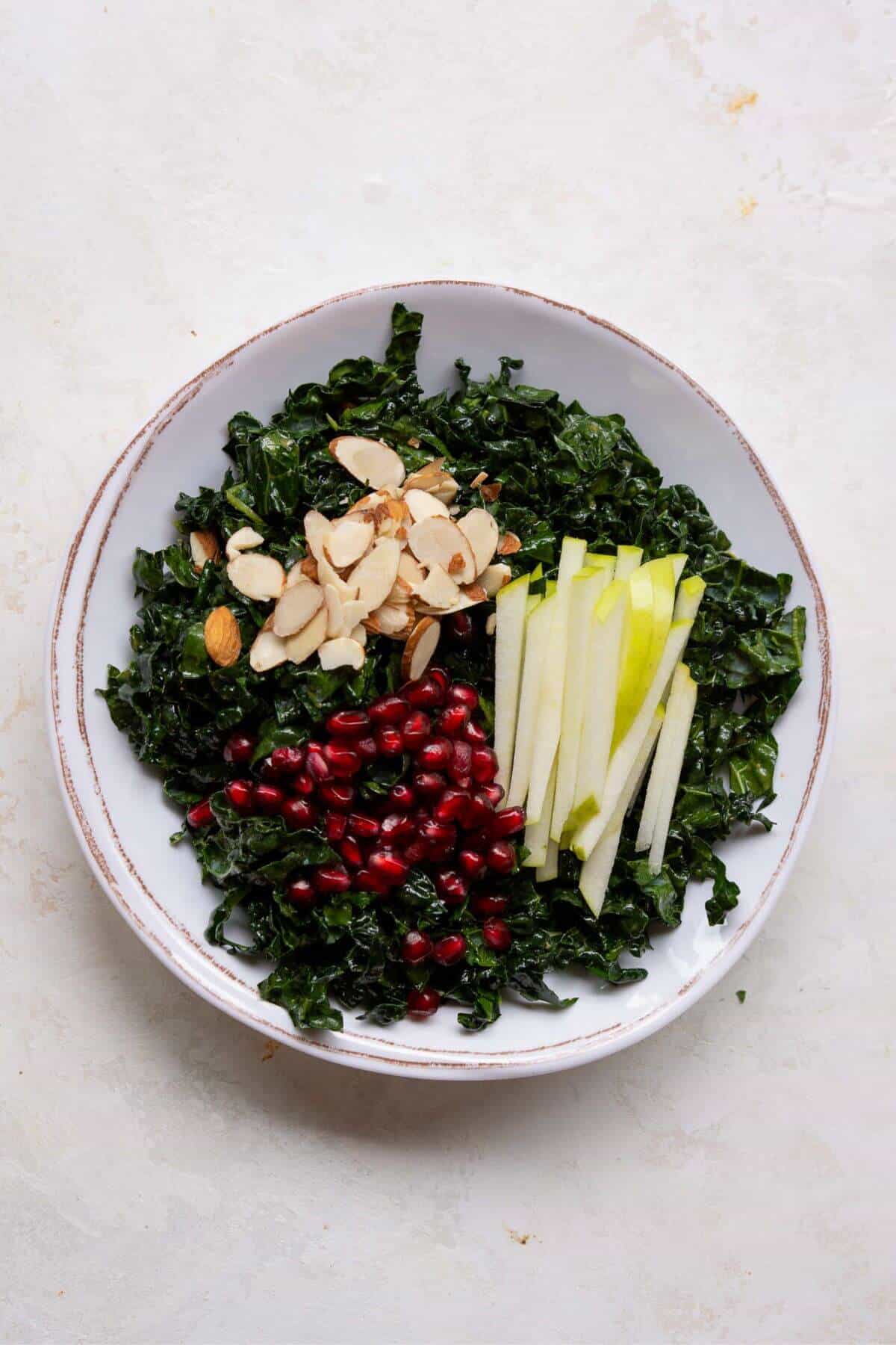Adding sliced almonds, fresh pomegranate seeds, and sliced green apple to messaged kale.