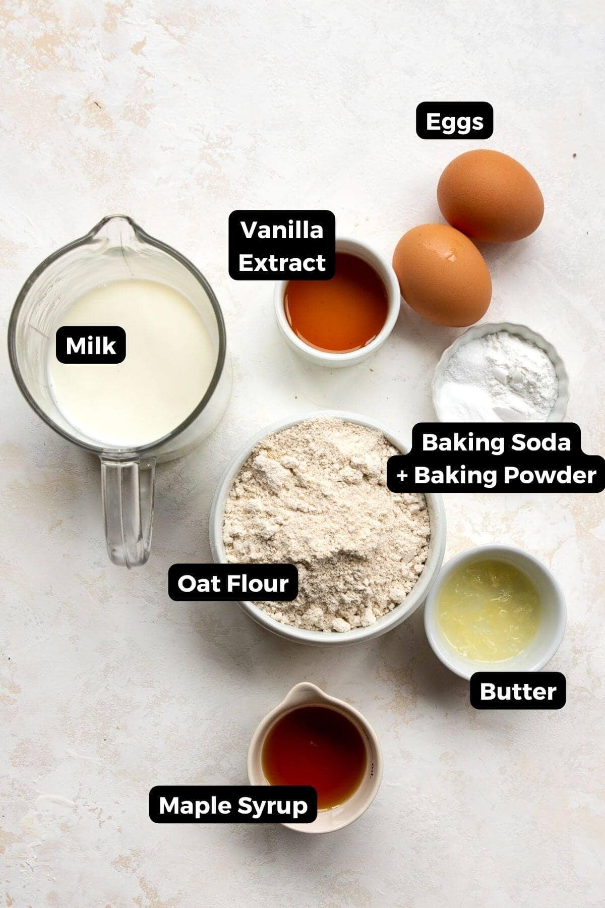 All ingredients in out flour pancakes in small bowls with labels, including: oat flour, milk, eggs, vanilla extract, baking soda, baking powder, butter, and maple syrup.