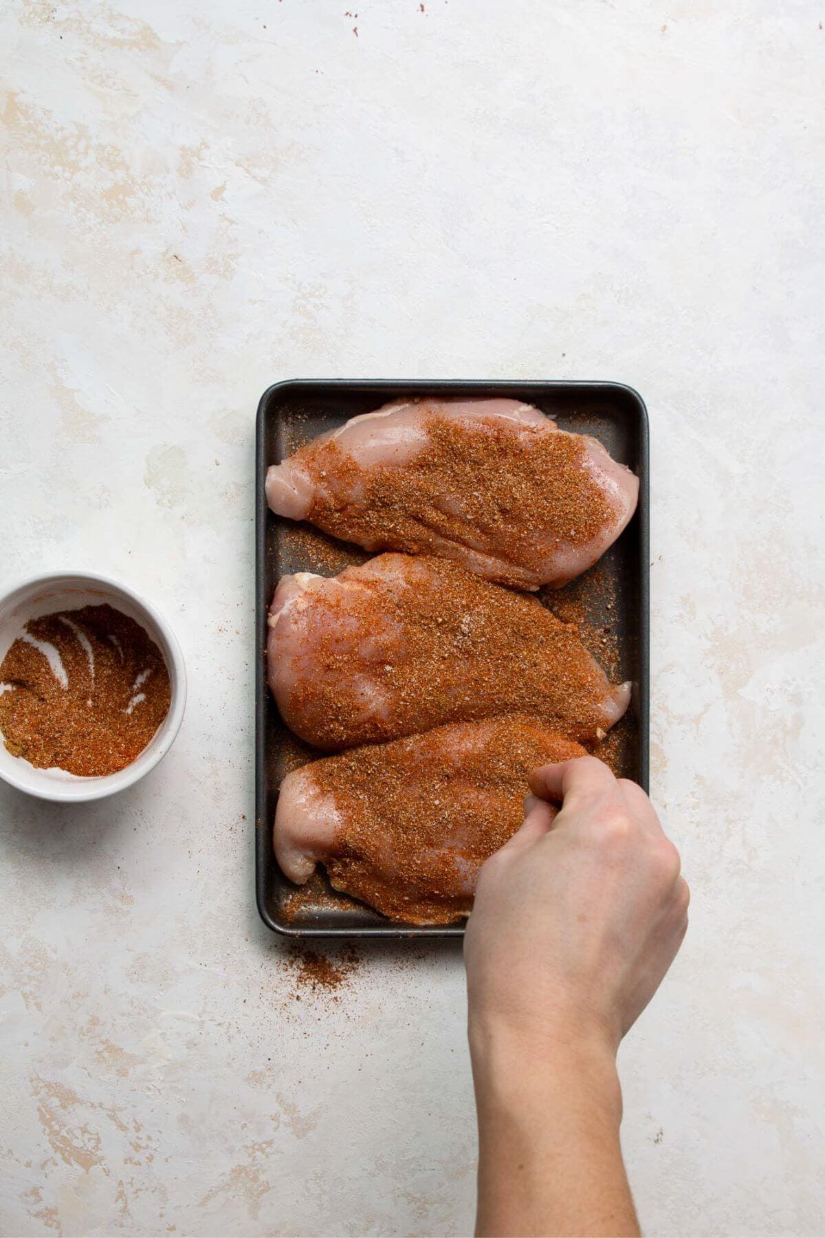 Hand seasoning raw chicken breasts with fajita spice on a sheet pan.