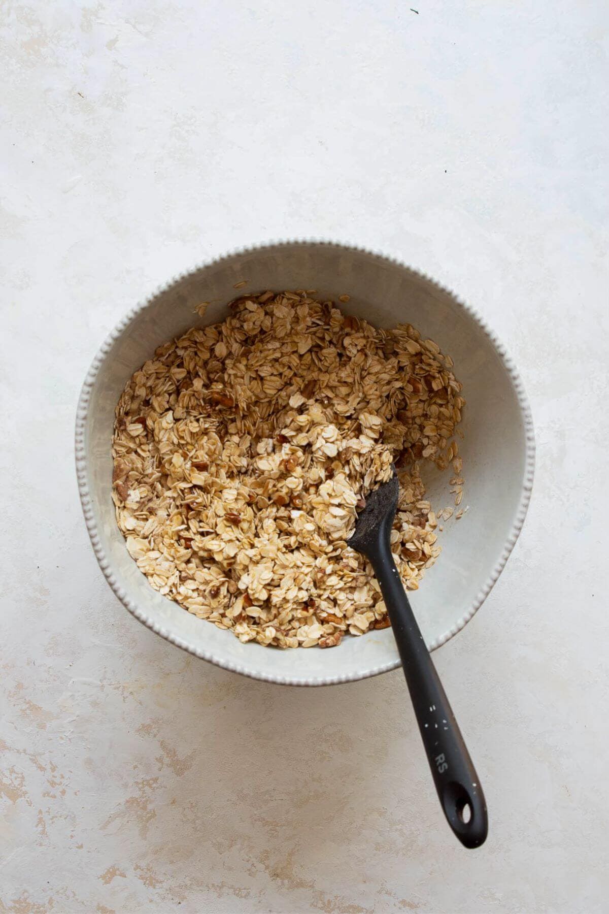 Healthy granola mixture tossed with melted coconut oil, maple syrup, and vanilla in a white bowl, ready to spread on a baking sheet.