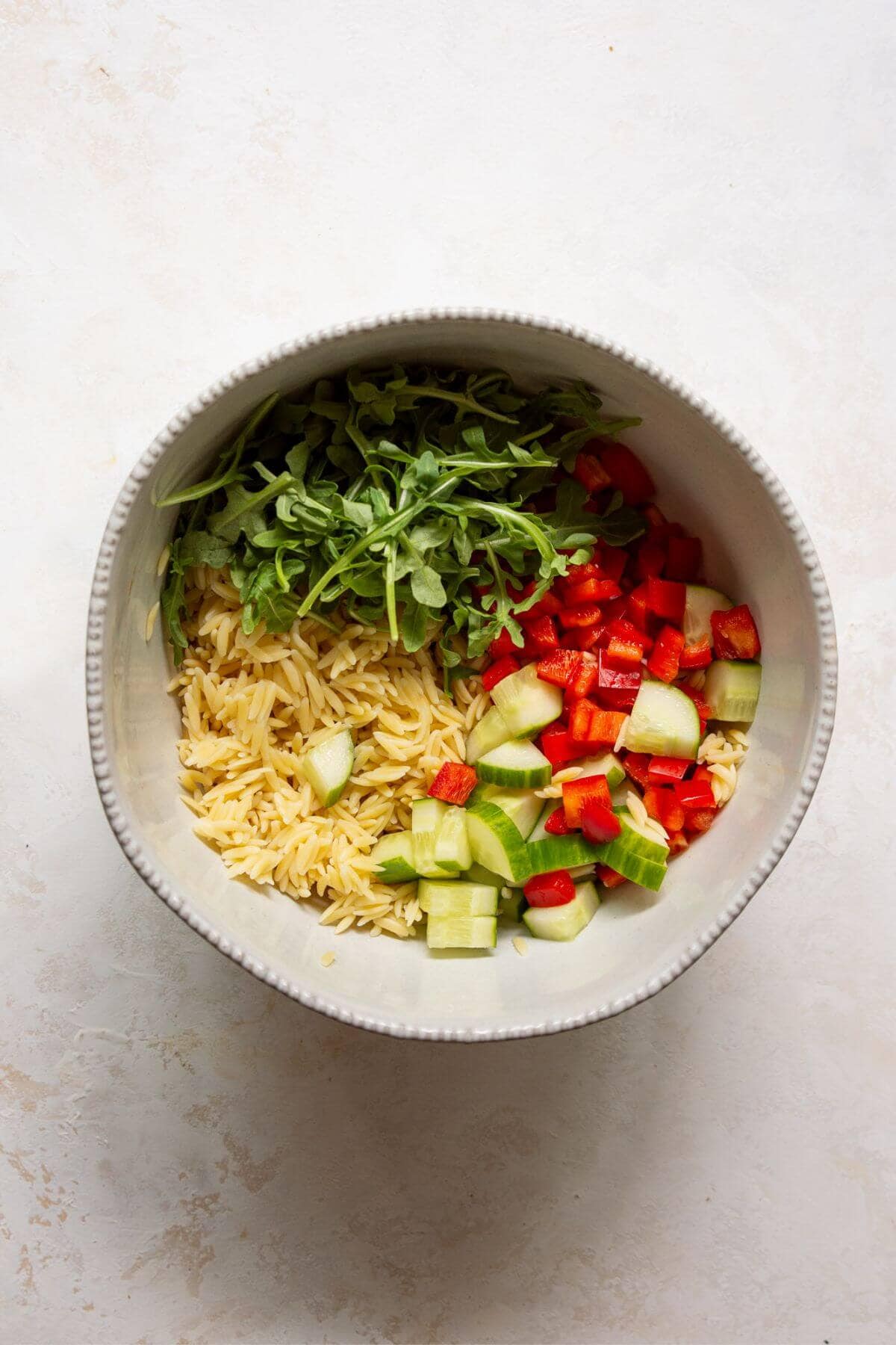 Cooked orzo in a bowl topped with arugula, diced cucumber, and red bell pepper before tossing.
