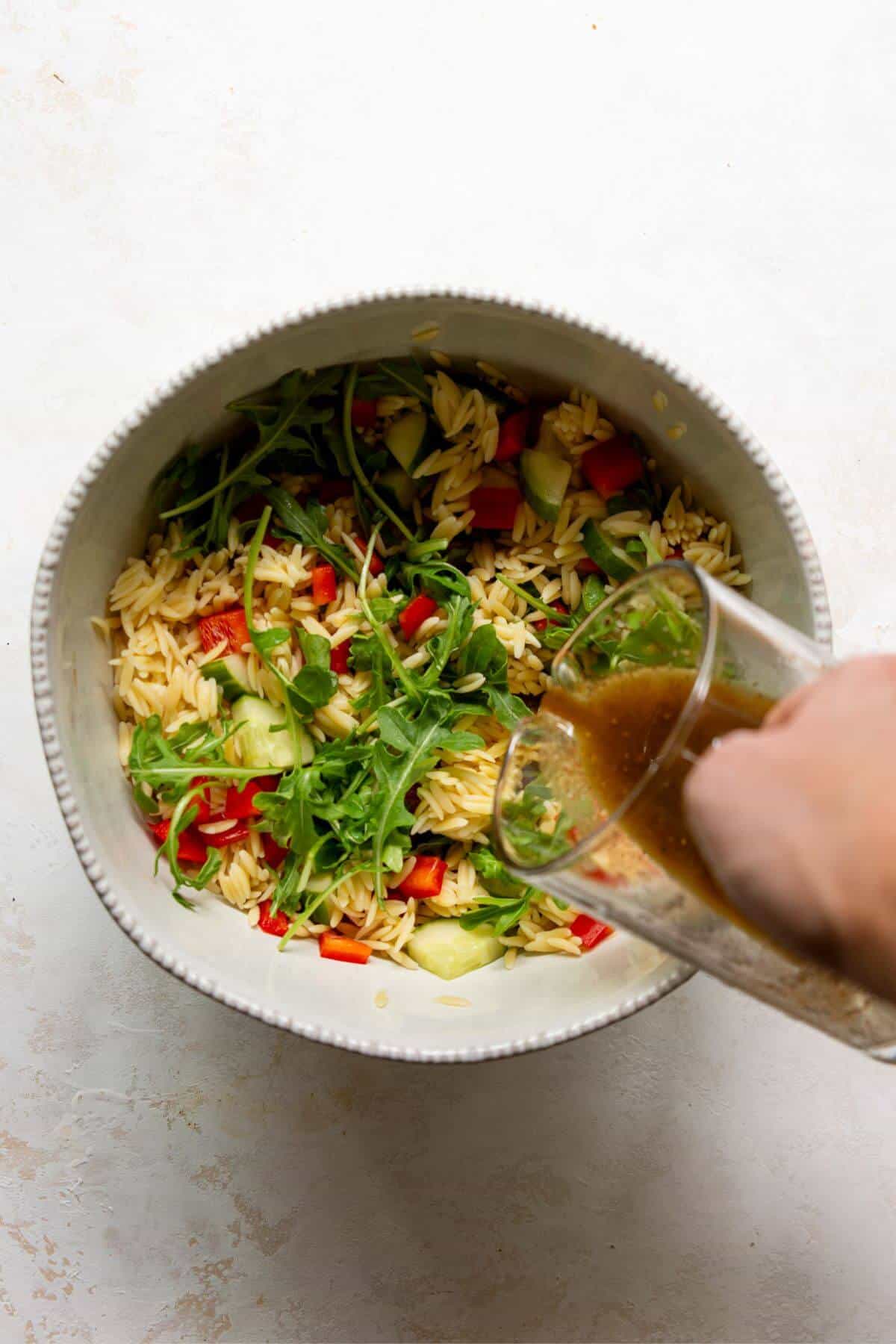 Balsamic dressing being poured over orzo and chopped vegetables in a large bowl.