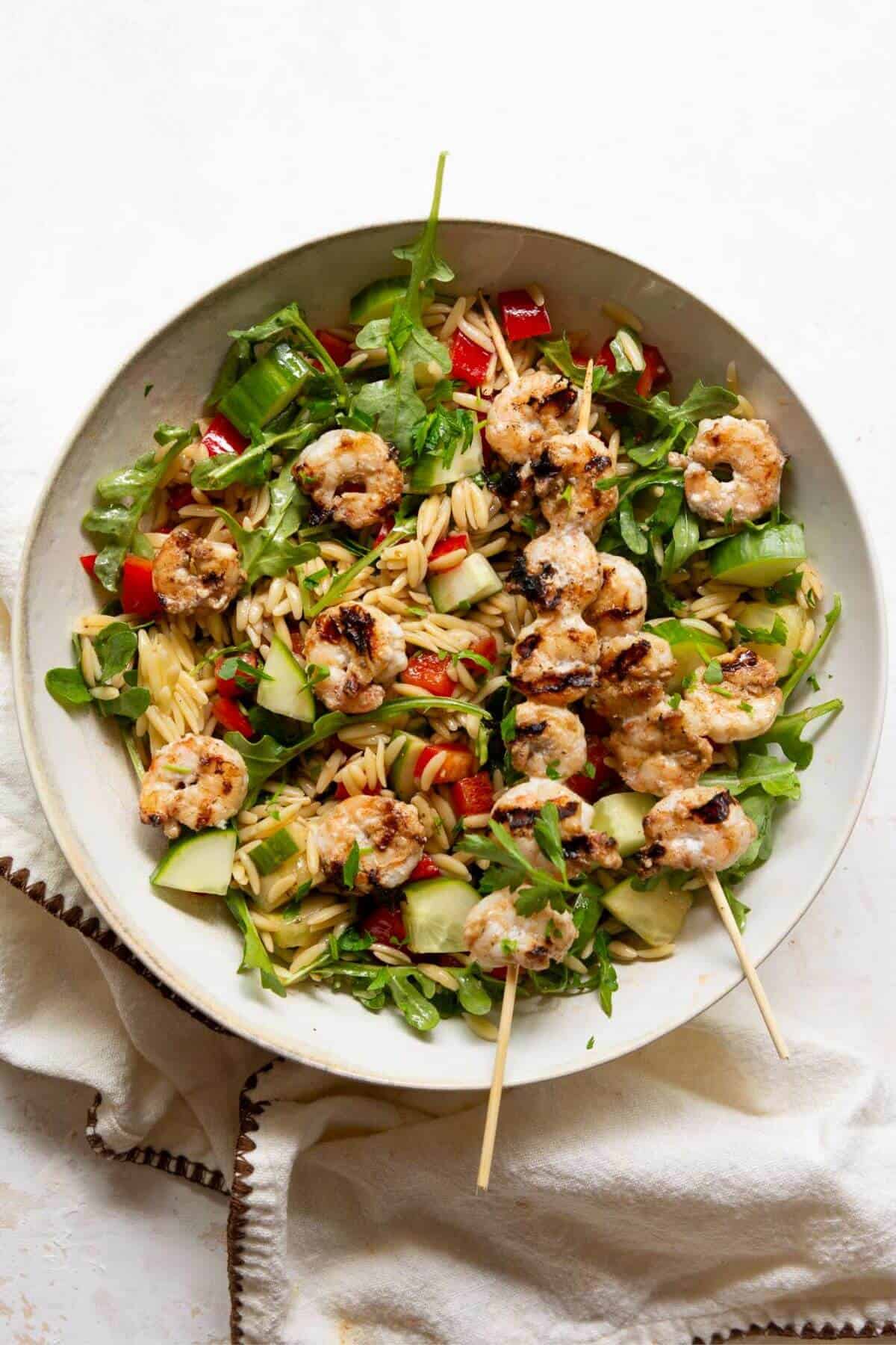 Finished grilled shrimp orzo salad plated with skewers, arugula, and fresh parsley.