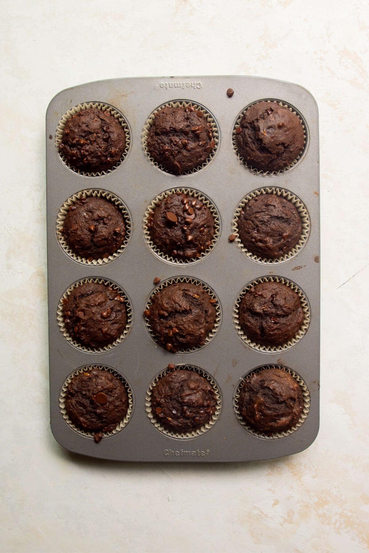 Twelve baked chocolate banana muffins with domed tops and chocolate chips in a muffin tin.