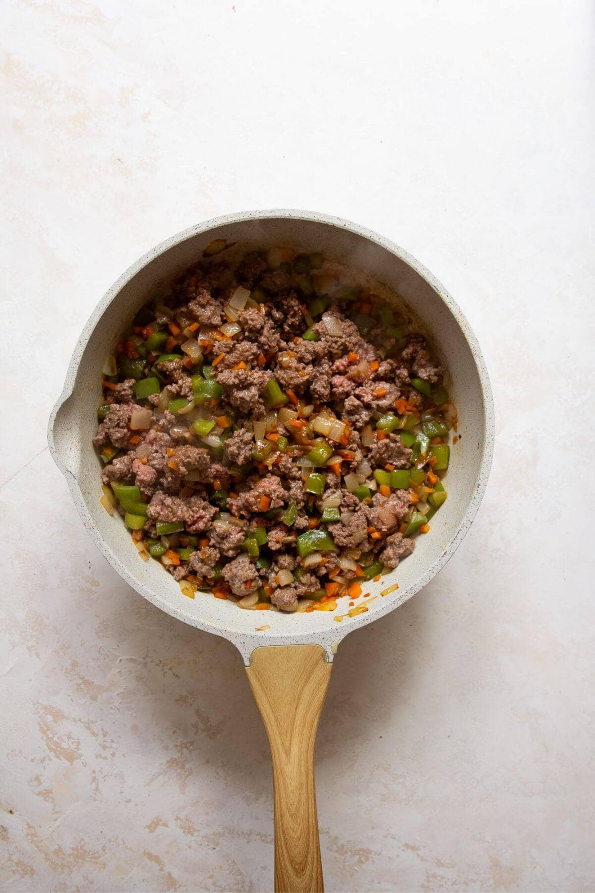 Ground beef browning in a skillet next to cooked vegetables for sloppy joes.