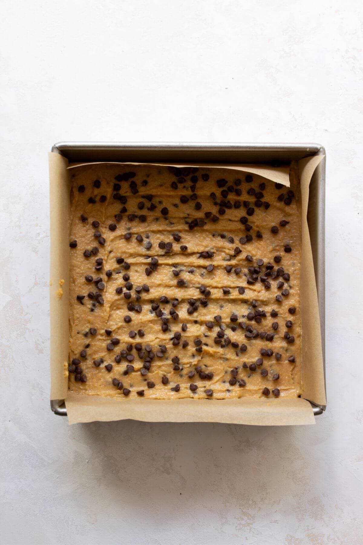 Chickpea blondie batter spread in a parchment-lined pan and topped with chocolate chips.