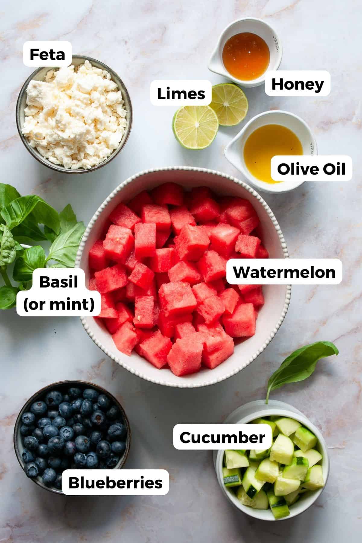 Watermelon blueberry salad ingredients in bowls: cubed watermelon, blueberries, cucumber, feta, basil, lime, honey, and olive oil.