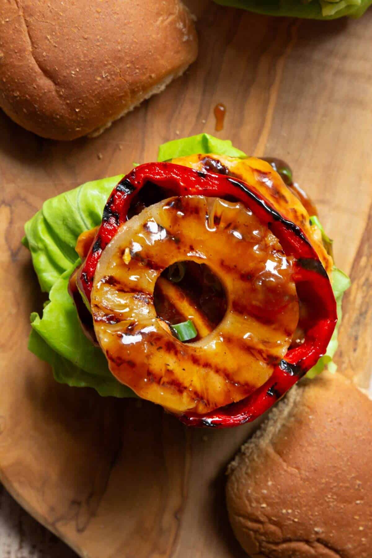 Assembling teriyaki chicken burgers with caramelized pineapple, lettuce, charred bell pepper, and extra teriyaki sauce.