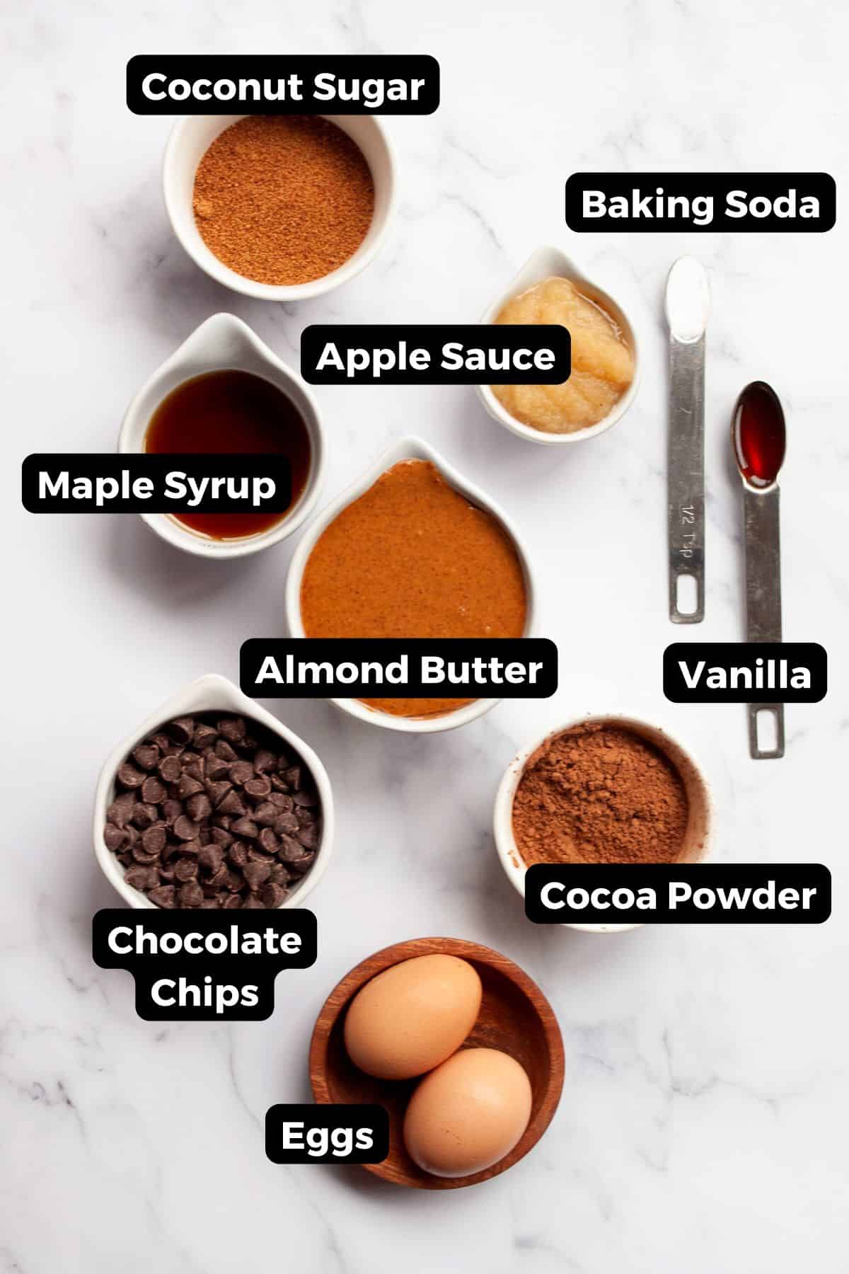 All ingredients for flourless brownies including almond butter, cocoa powder, maple syrup, eggs, and coconut sugar.