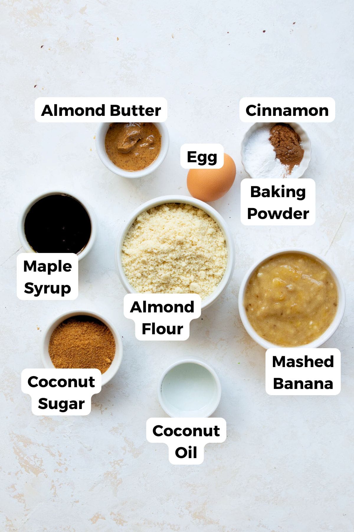 Ingredients for healthy banana bread donuts including almond flour, mashed banana, almond butter, maple syrup, coconut sugar, coconut oil, egg, and cinnamon.