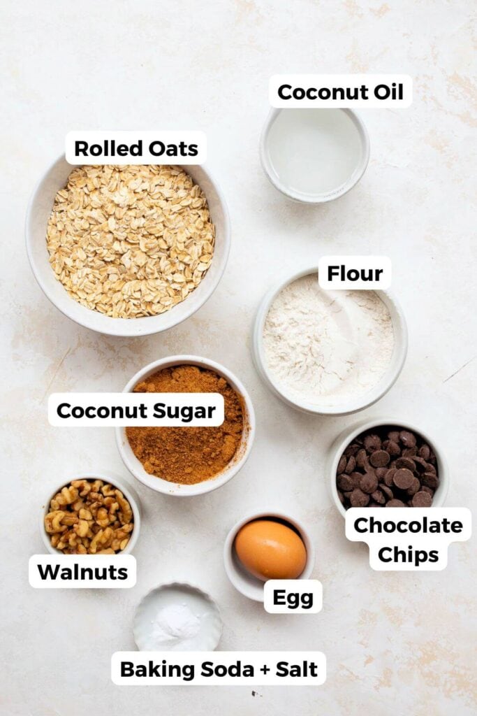 the ingredients in healthy oatmeal walnut cookies including rolled oats, coconut oil, gluten-free flour, coconut sugar, an egg, chocolate chips, walnuts, and baking soda