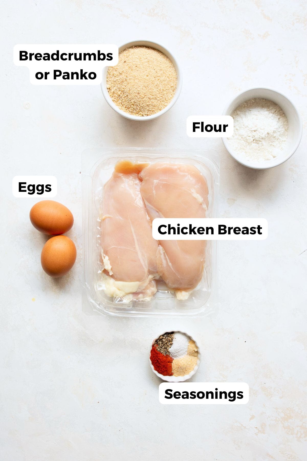 All the ingredients in air fryer chicken cutlets in small bowls including raw chicken breasts, flour, eggs, panko breadcrumbs, and seasonings.