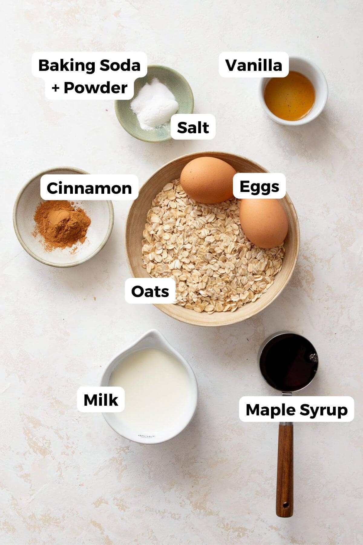 ingredients in banana oat blender muffins in bowls including rolled oats, eggs, milk, maple syrup, vanilla, salt, baking soda, baking powder, and cinnamon