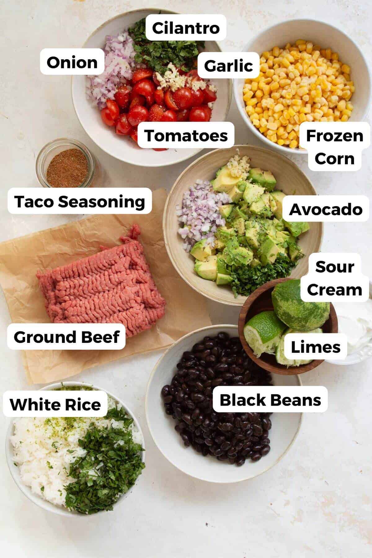 all ingredients in ground beef burrito bowls in small bowls including frozen corn, tomatoes, onion, cilantro, avocado, limes, black beans, ground beef, taco seasoning, and white rice