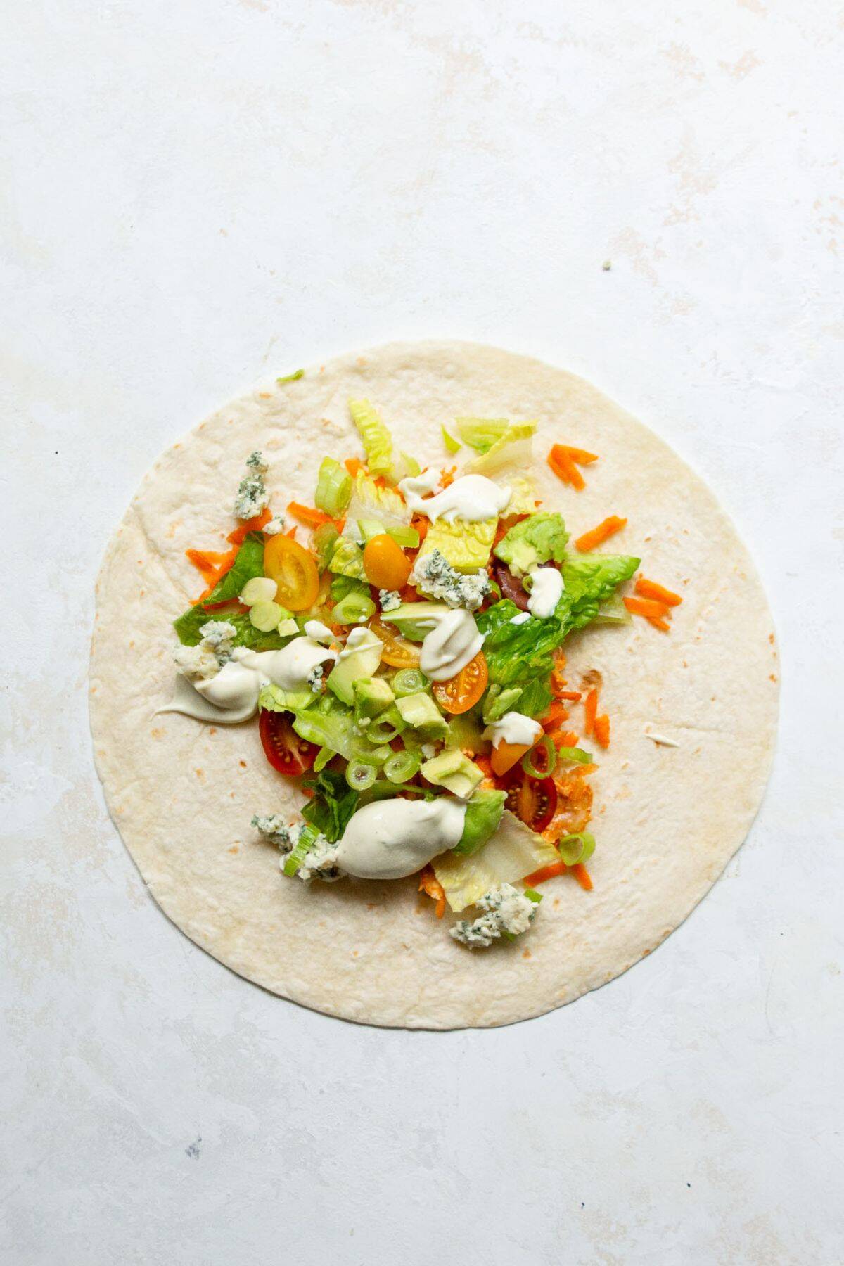 Filling a tortilla with cooked buffalo chicken, chopped lettuce, tomatoes, carrots, green onion, blue cheese, avocado, and caesar dressing before wrapping.