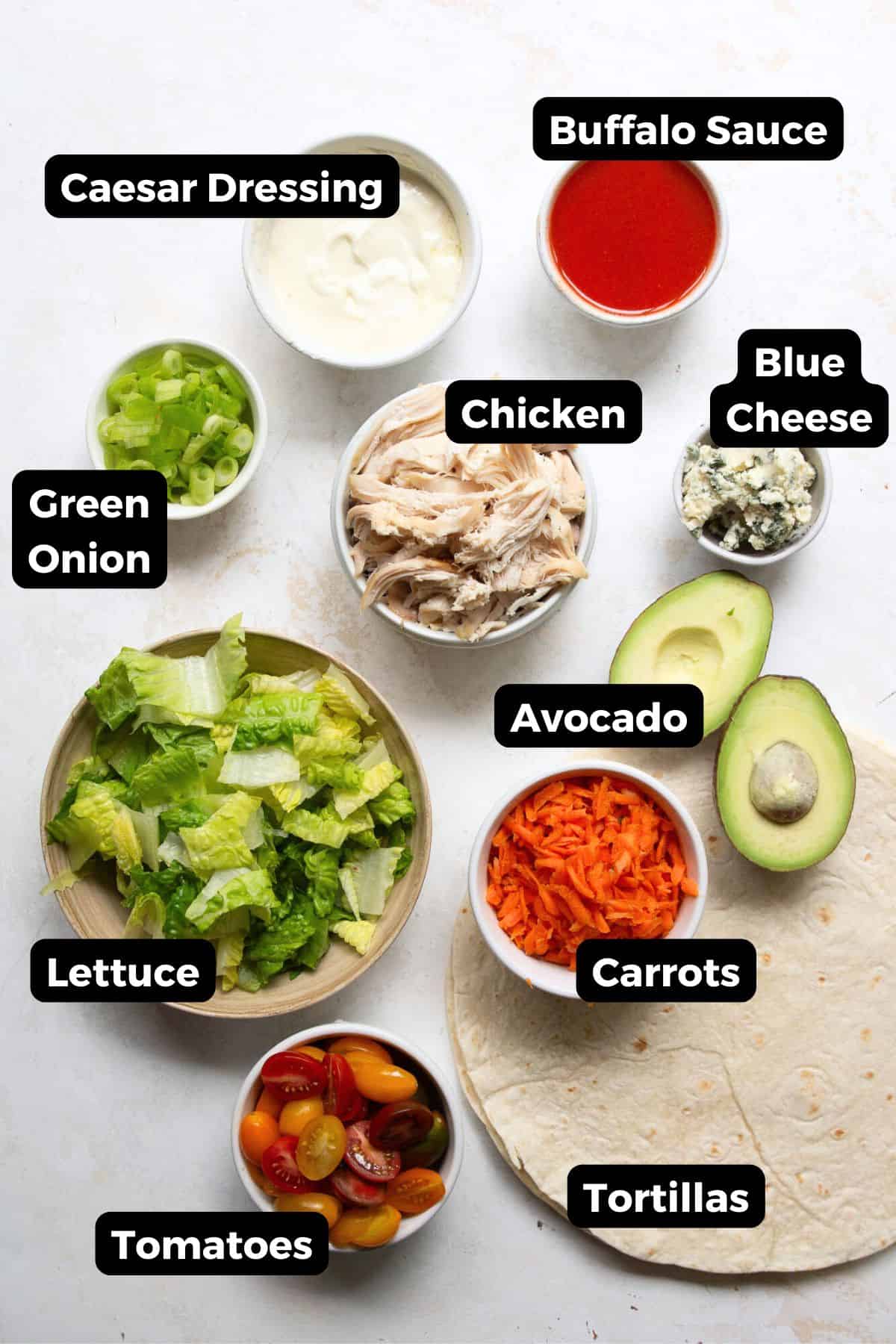 All ingredients in buffalo chicken caesar wraps in small bowls with labels, including: tortillas, rotisserie chicken, green onion, buffalo sauce, greek yogurt caesar dressing, avocado, carrots, tomatoes, and lettuce.