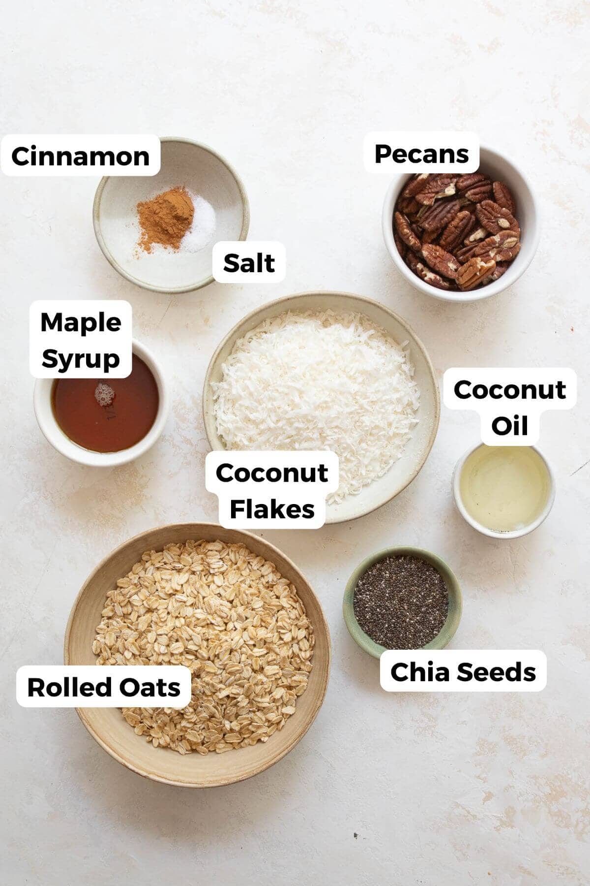 all ingredients in coconut granola in bowls including rolled oats, coconut flakes, coconut oil, chia seeds, pecans, cinnamon, maple syrup, and salt