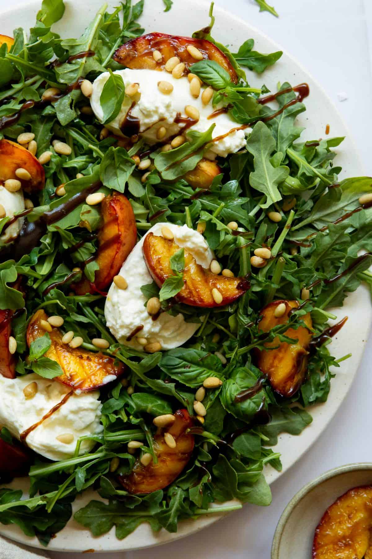 Grilled peach and burrata salad served on a platter with arugula, fresh basil, toasted pine nuts, and a drizzle of balsamic vinaigrette.