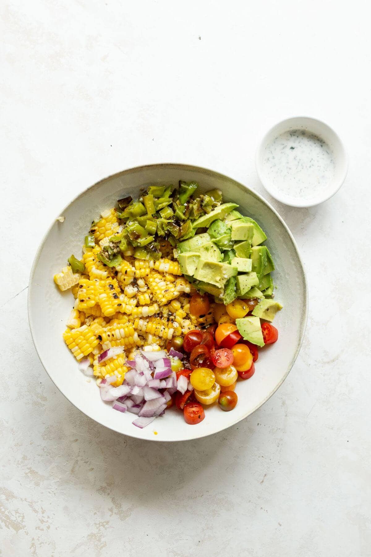 Grilled corn cut off the cobb, shishito peppers, tomatoes, avocado, and red onion in a bowl before tossing.