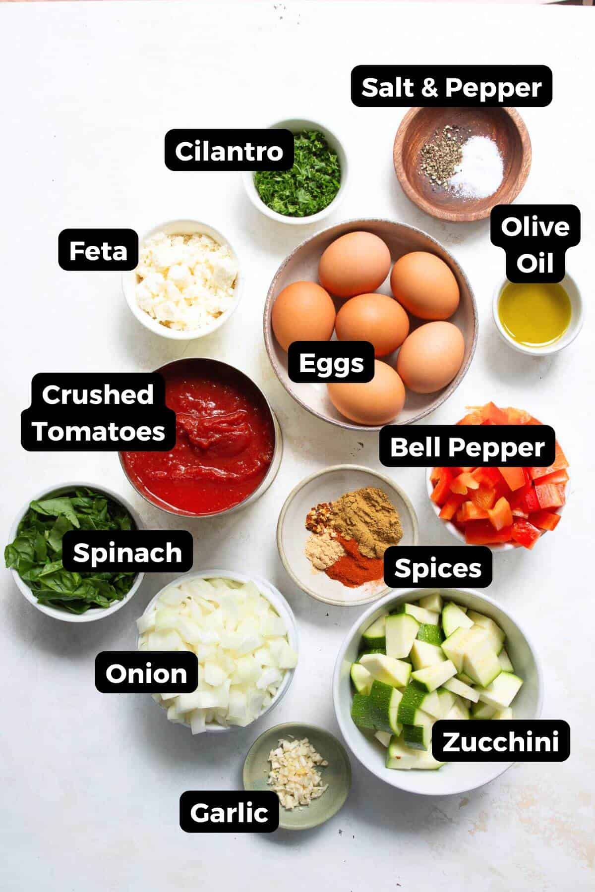 All ingredients needed to make Mediterranean shakshuka with feta, labeled and ready to cook-including eggs, zucchini, red bell pepper, yellow onion, garlic, spinach, cilantro, crushed tomatoes, cumin, paprika, ground ginger, red chili flakes, olive oil, and crumbled feta.