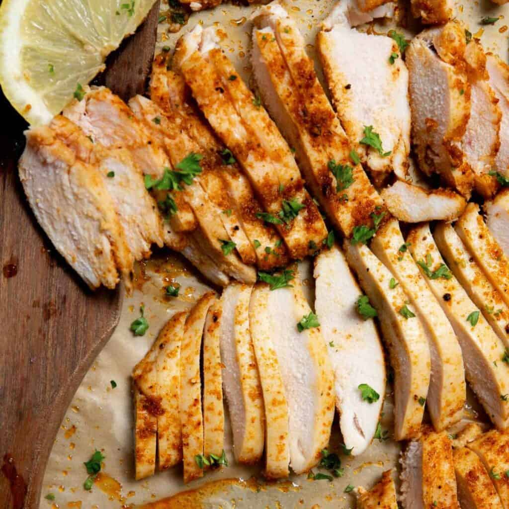 Juicy strips of oven-baked thin sliced chicken breast with a golden brown crust, seasoned with garlic, onion, and paprika, garnished with fresh lemon and parsley.