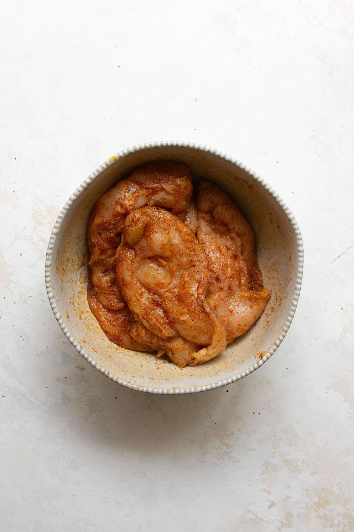 Raw thin-sliced chicken breast in a bowl, tossed with olive oil and seasonings like garlic powder, onion powder, and paprika.