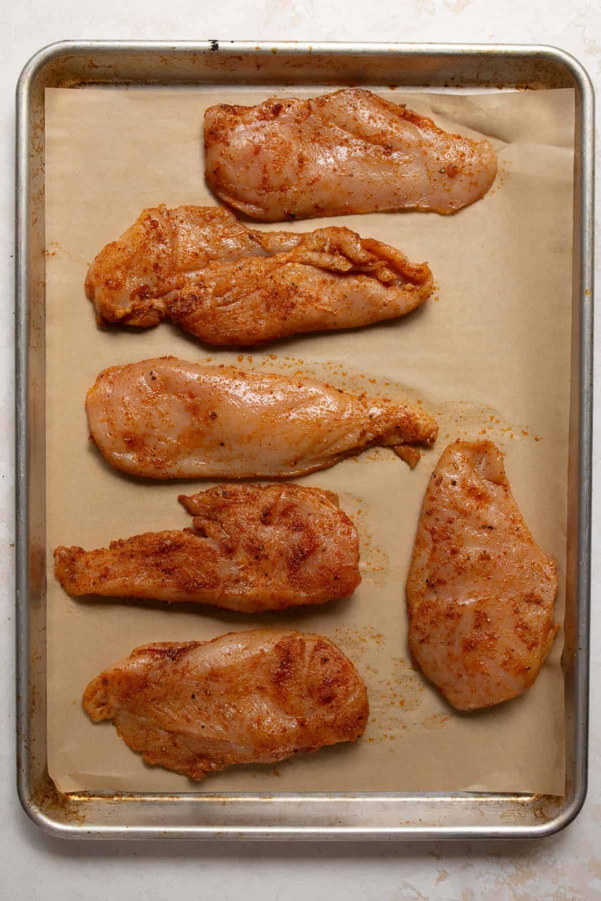 Raw thin-sliced chicken breast arranged on a baking sheet, ready to be baked with seasoning.