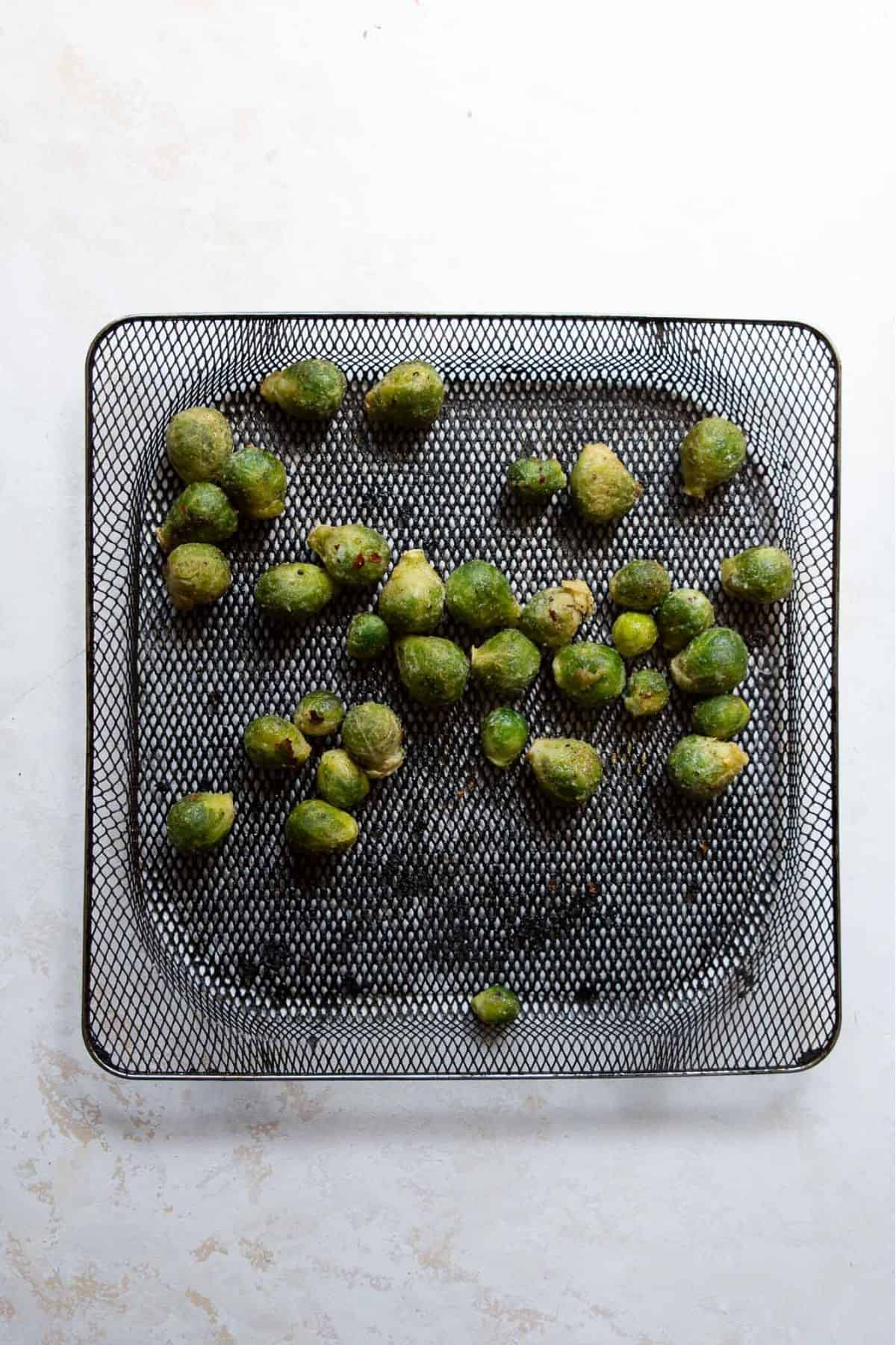 Seasoned frozen Brussel sprouts spread out in an air fryer basket before cooking.