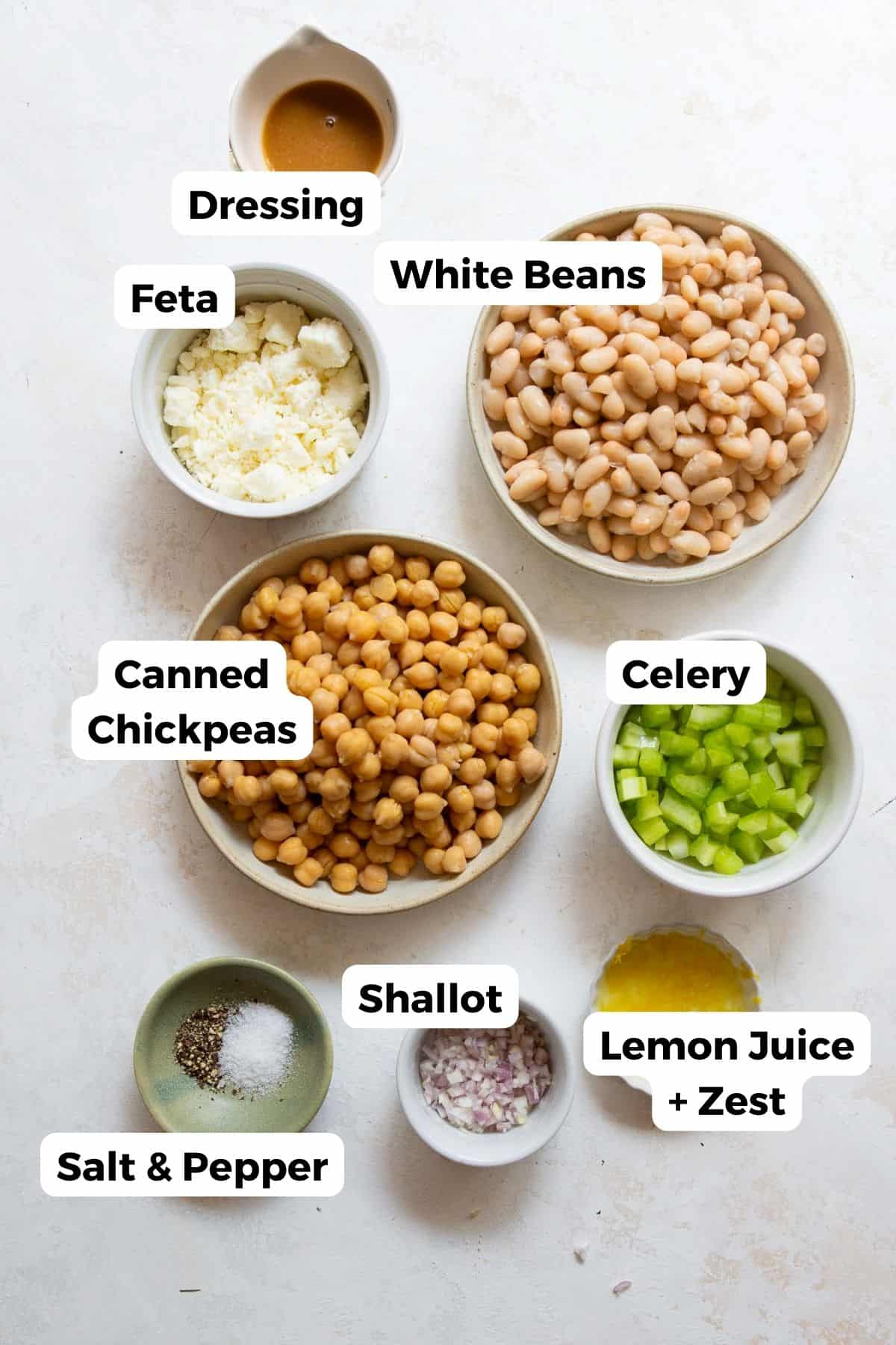 All ingredients in Mediterranean dense bean salad in labeled bowls, including: canned chickpeas, white beans, lemon zest, dill, shallots, celery, salt, pepper, feta, and a tangy Dijon-honey vinaigrette.