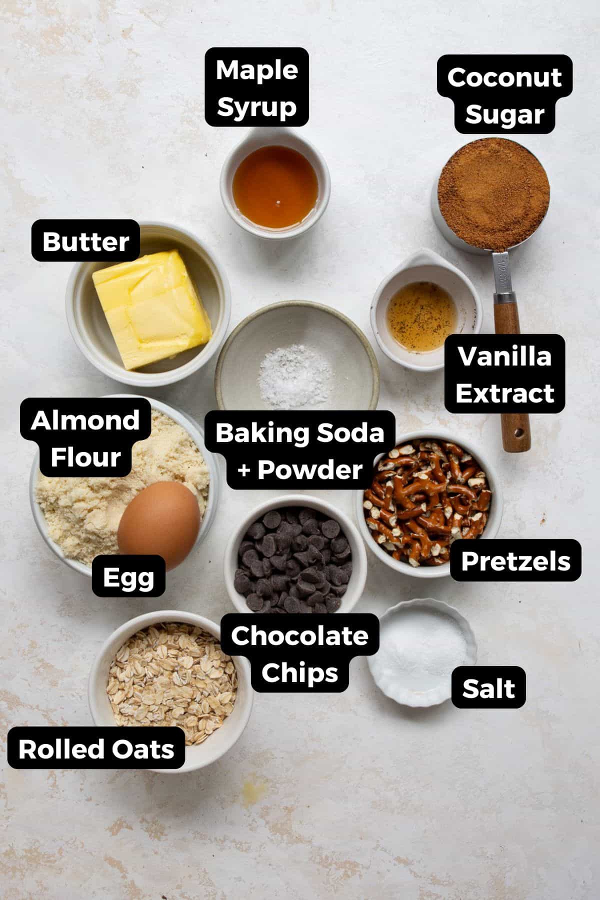 All ingredients for healthy cowboy pretzel cookies labeled: almond flour, rolled oats, coconut sugar, maple syrup, butter, egg, vanilla, baking soda, salt, chocolate chips, pretzels, flaky sea salt.