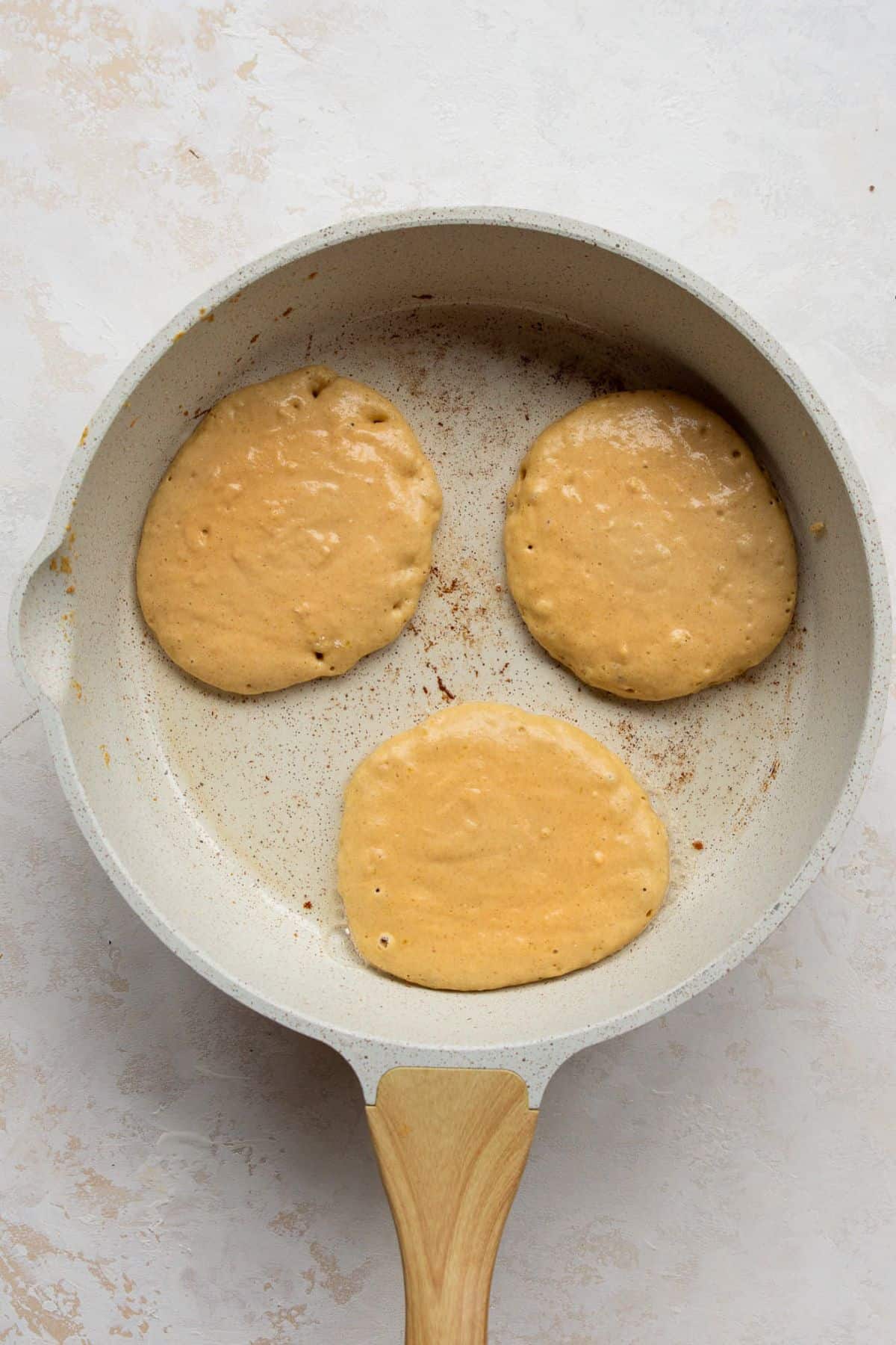 Three pumpkin protein pancakes cooking in a greased non-stick pan on the stove.
