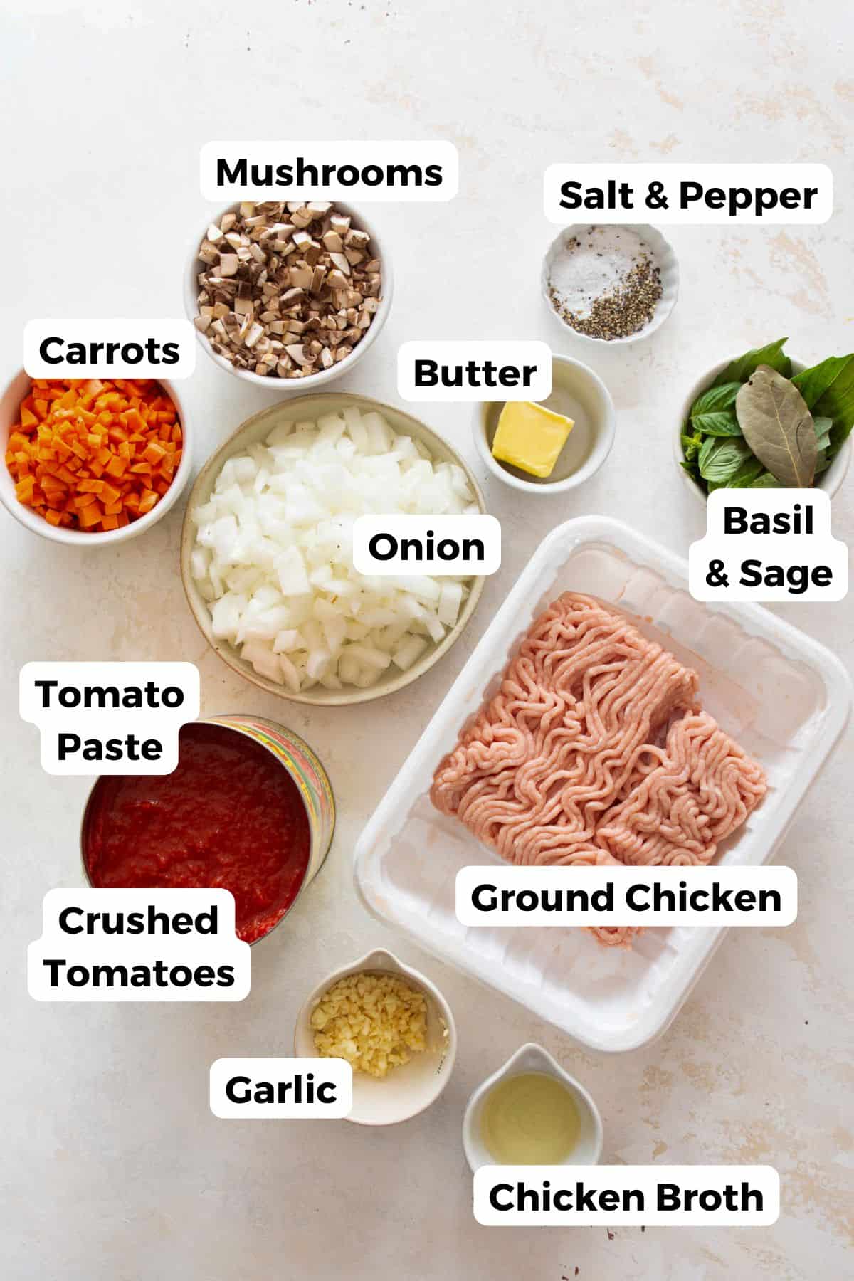 All ingredients in chicken bolognese recipe in bowls labeled, including: ground chicken, carrots, onions, mushrooms, garlic, crushed tomatoes, tomato paste, basil, bay leaves, and olive oil.