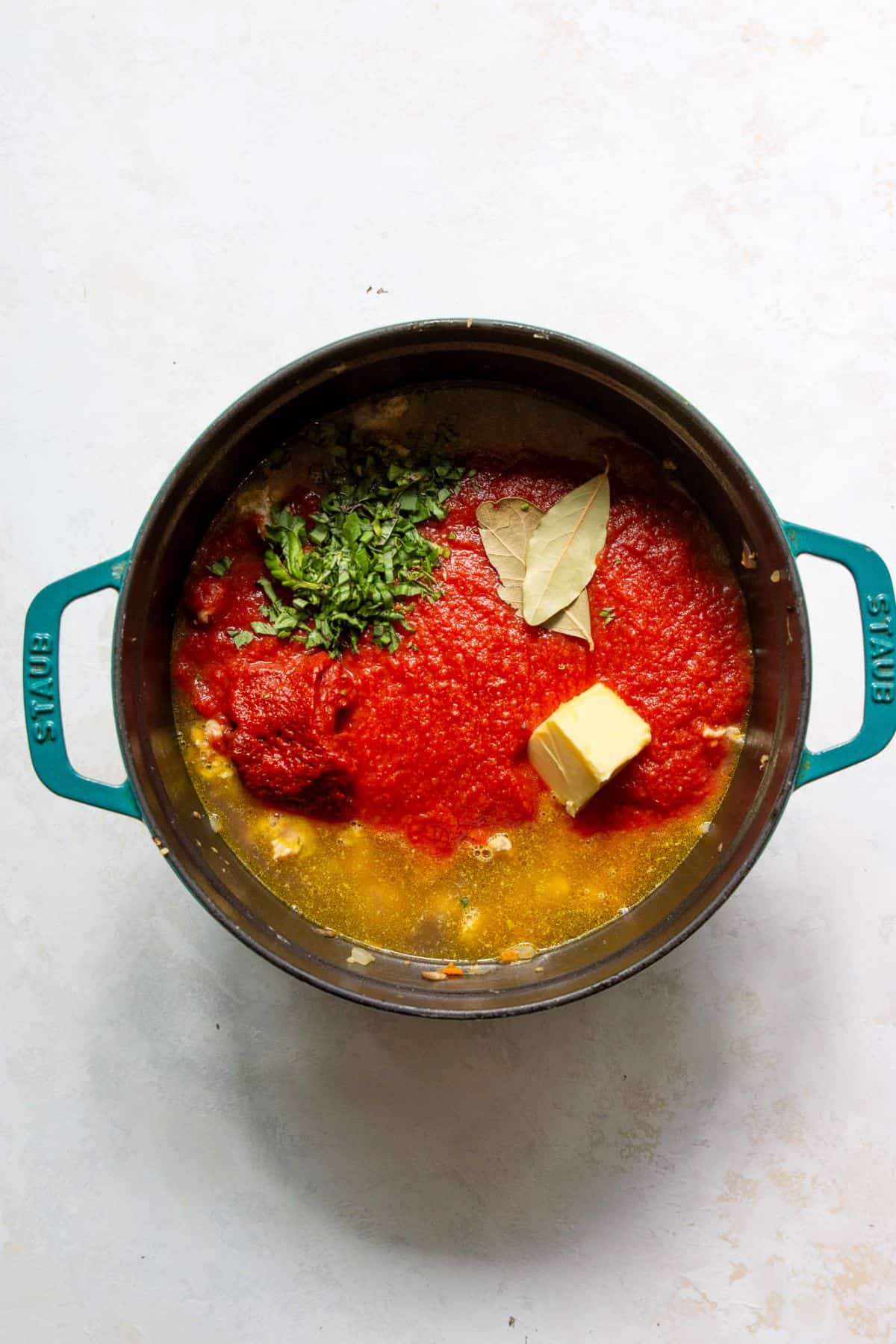 Pouring crushed tomatoes, tomato paste, fresh basil, bay leaves, and butter into sautéed veggies and ground chicken to make the chicken bolognese sauce.