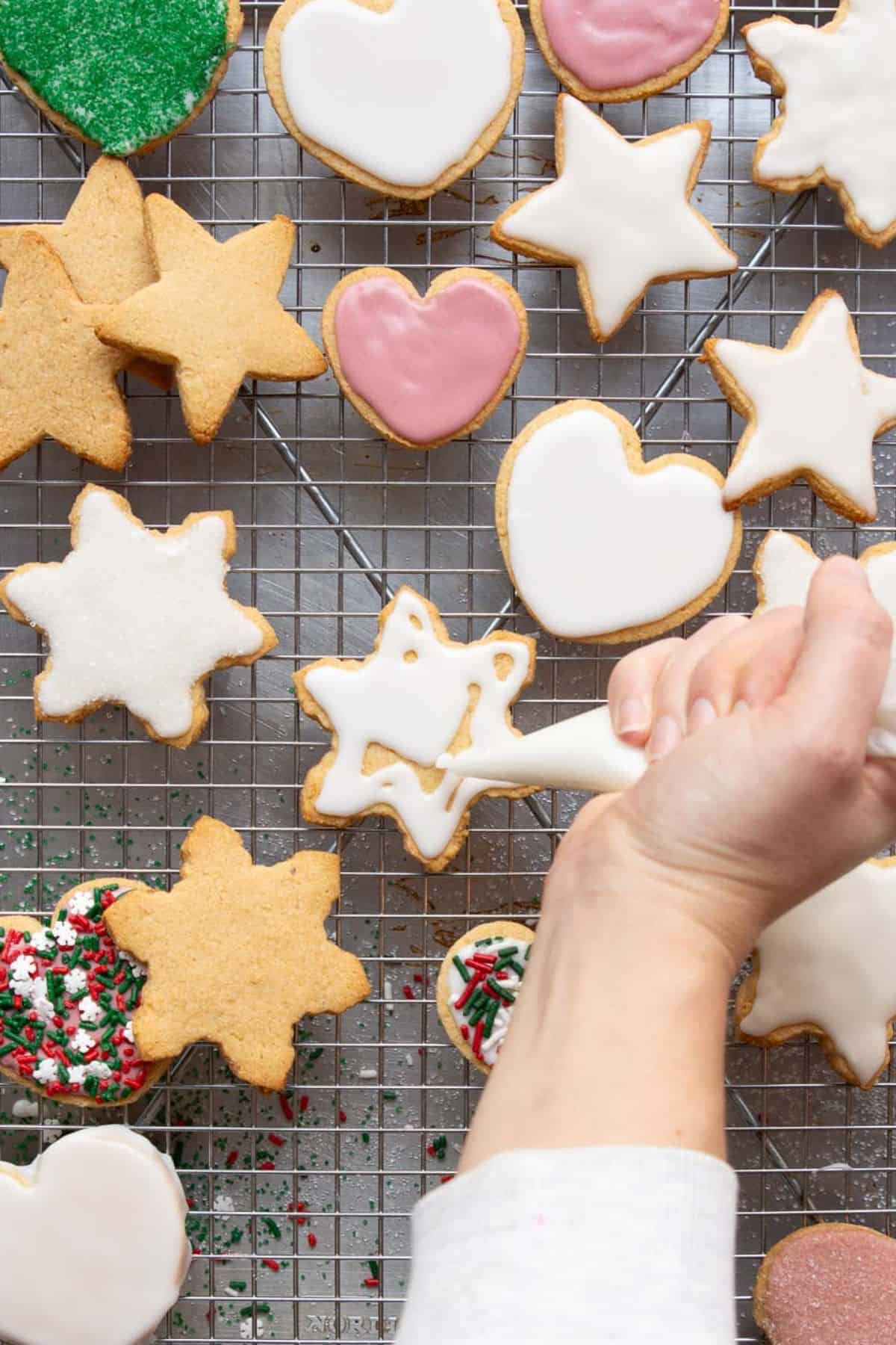 Hand piping white icing onto almond flour sugar cookies shaped like stars and hearts on a wire cooling rack.