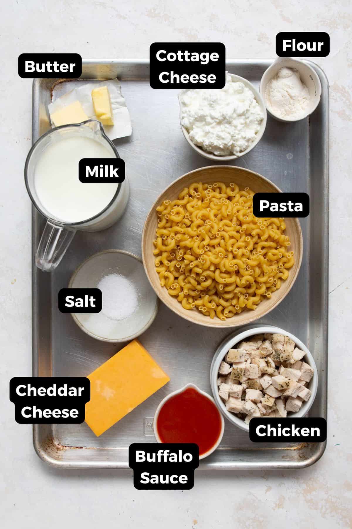 All ingredients in high-protein buffalo chicken mac and cheese in small bowls, including: elbow pasta, milk, cottage cheese, flour, milk, salt, chicken, cheddar cheese, and buffalo sauce.