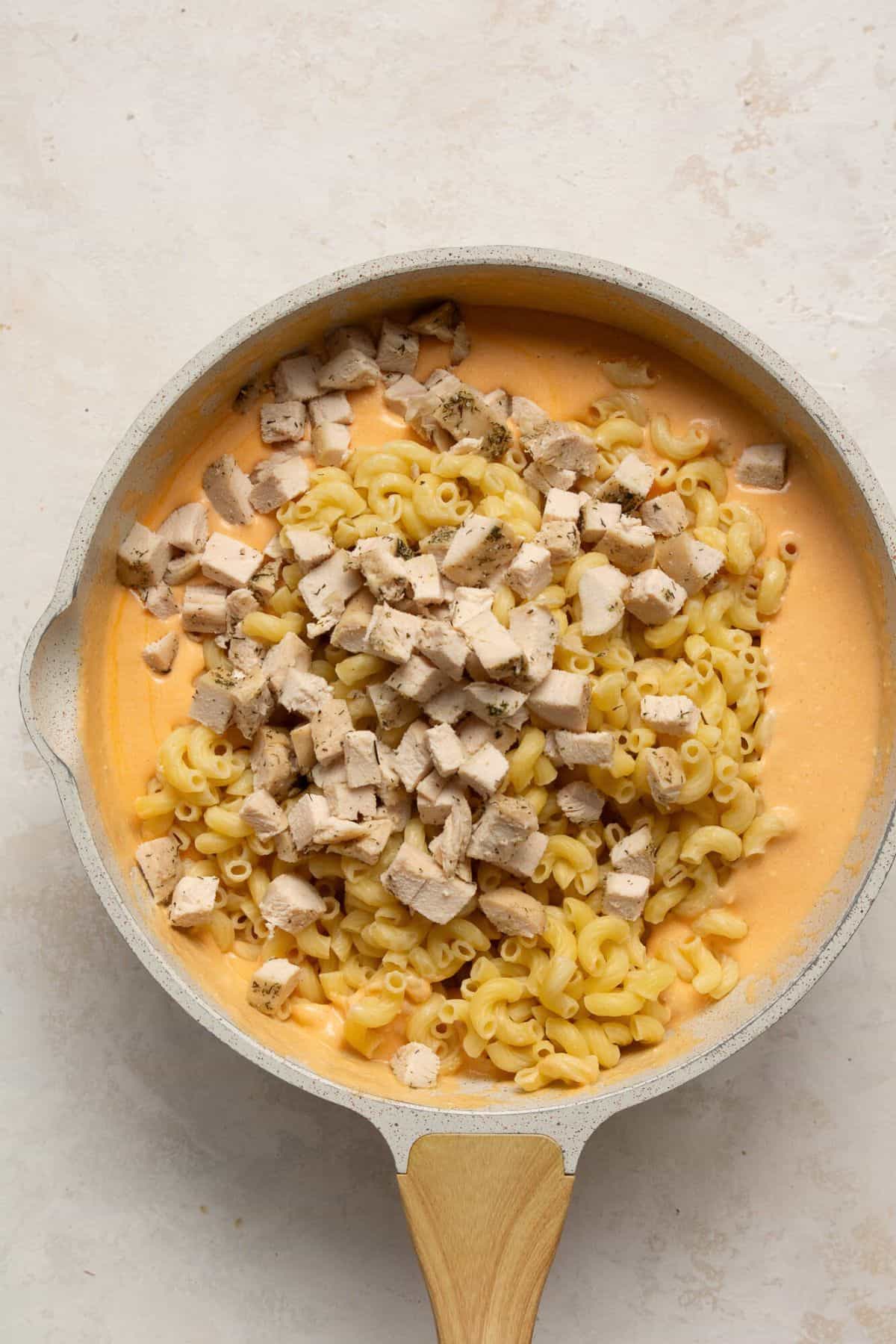 Adding cooked elbow pasta and cubed chicken breasts to the mac and cheese sauce.