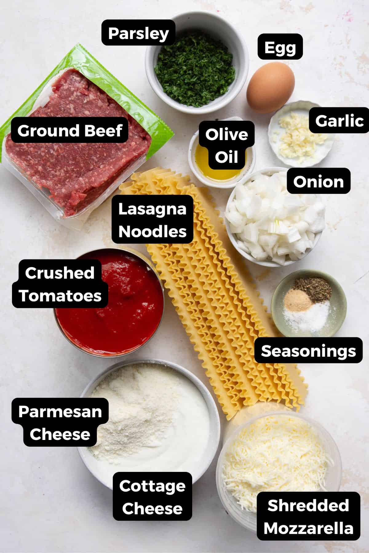 Ingredients for healthy cottage cheese lasagna labeled in bowls, including ground beef, cottage cheese, onion, tomatoes, noodles, and mozzarella.