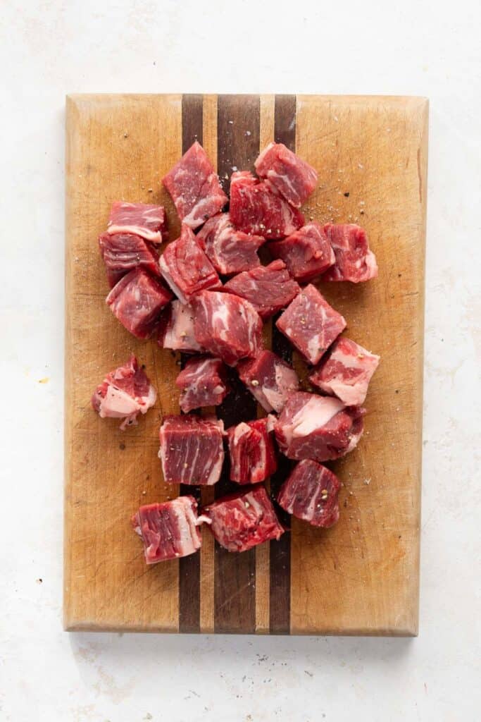 Raw sirloin steak cut into 1-inch cubes and seasoned with salt and pepper on a wooden cutting board.