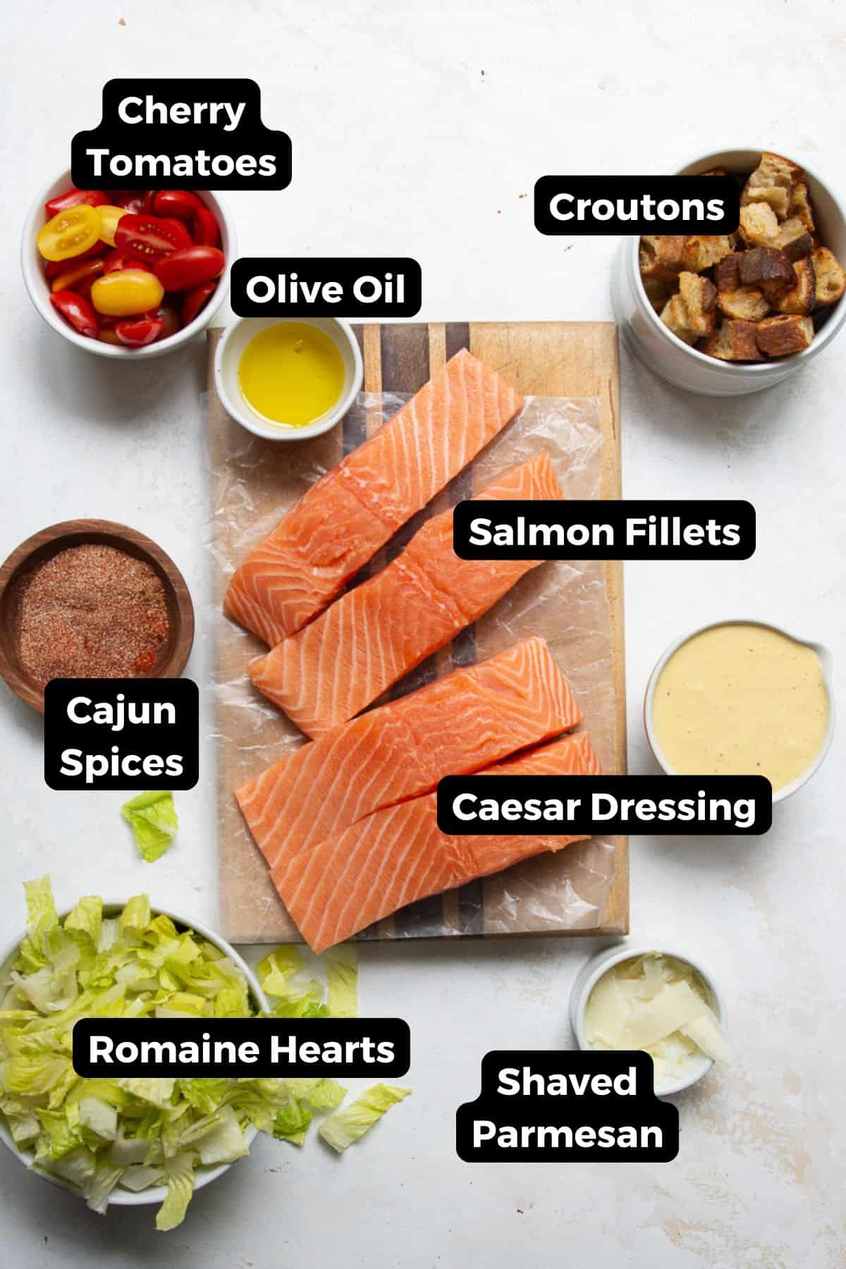 Ingredients for blackened salmon Caesar salad labeled, including: salmon fillets, Greek yogurt caesar dressing, Parmesan, romaine, cherry tomatoes, sourdough croutons, and Cajun blackening spices.