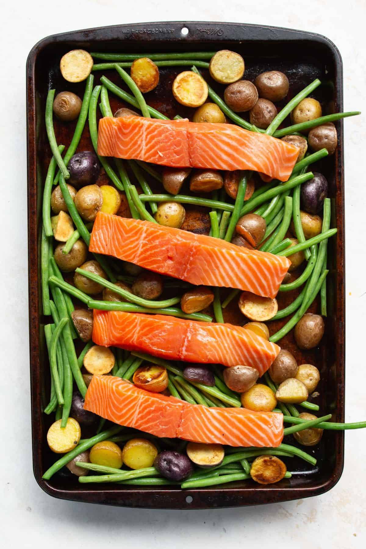 Raw salmon fillets placed on a sheet pan with roasted vegetables.