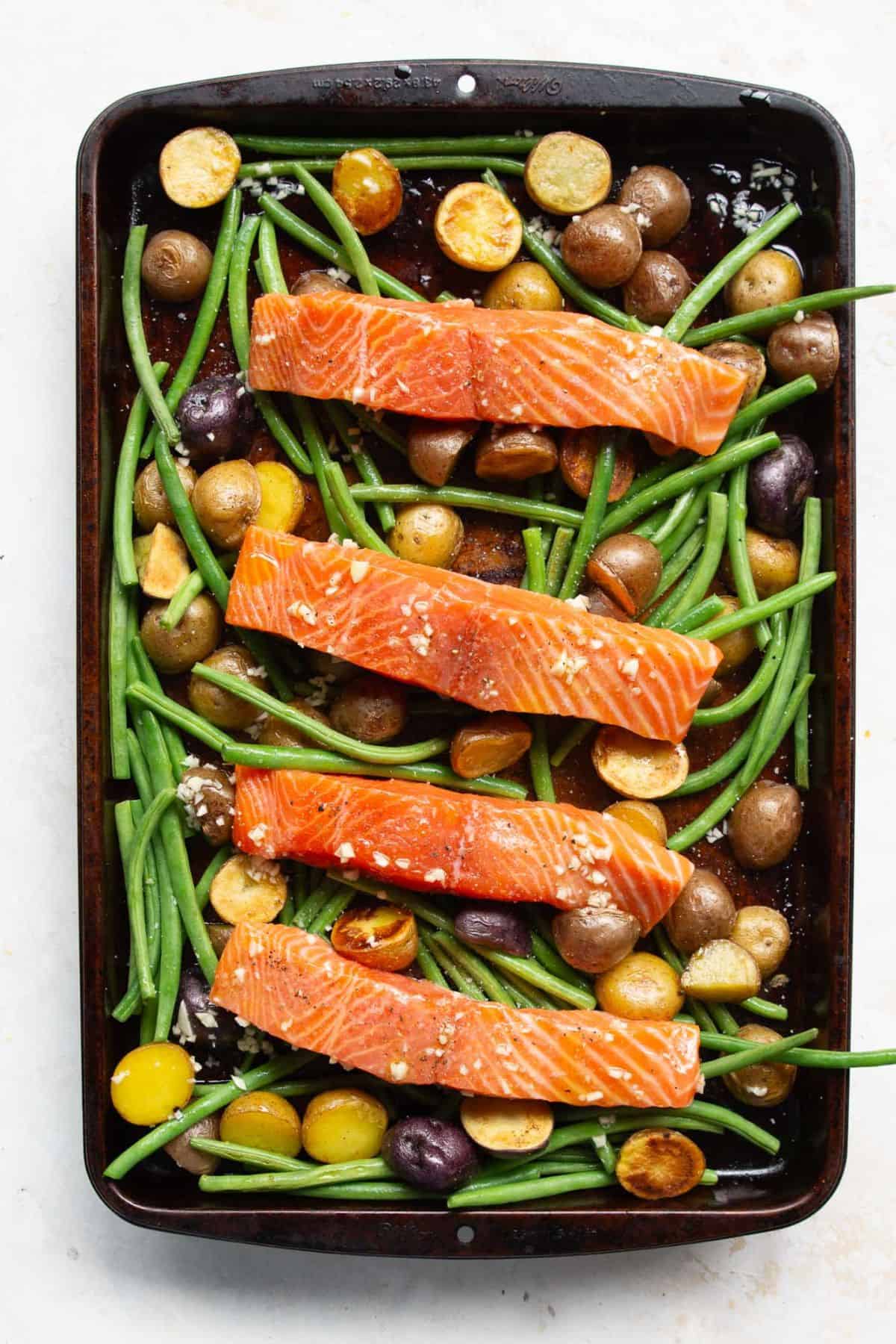 Raw seasoned salmon on top of roasted vegetables and potatoes on a sheet pan before roasting.