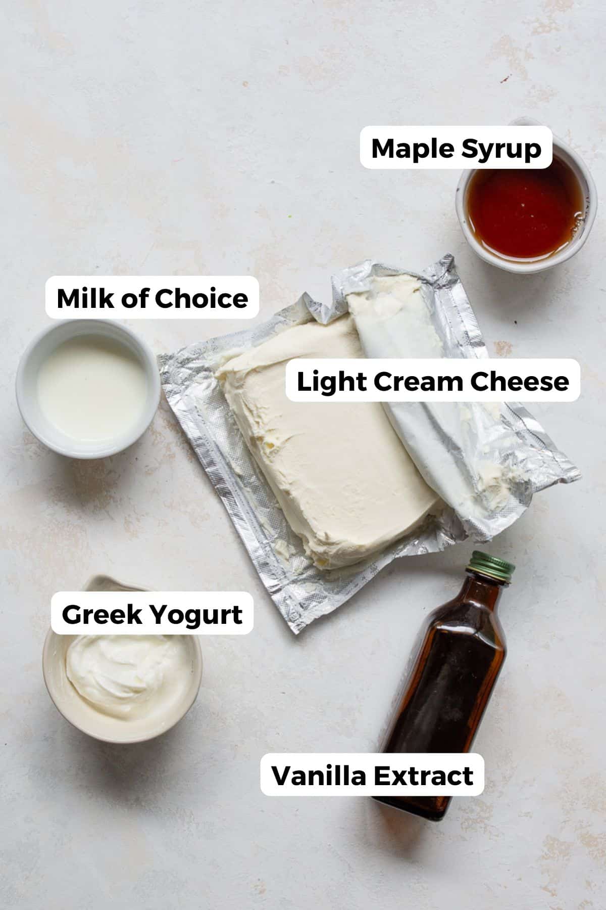 Ingredients for healthy cream cheese frosting laid out on a marble surface: light cream cheese, plain Greek yogurt, maple syrup, milk, and vanilla extract.